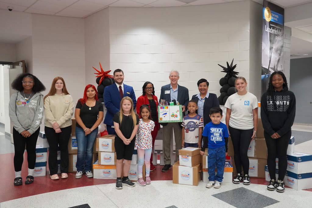 A group of students and adults pose together in a school hallway in front of stacks of donated supply boxes. One adult holds a decorated box of supplies while others smile beside them.