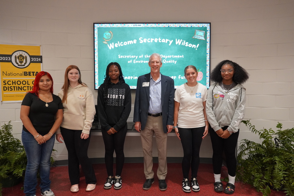 Five students stand with a man in front of a digital display that reads “Welcome Secretary Wilson! Secretary of the Department of Environmental Quality.” A National Beta School of Distinction banner hangs on the wall.