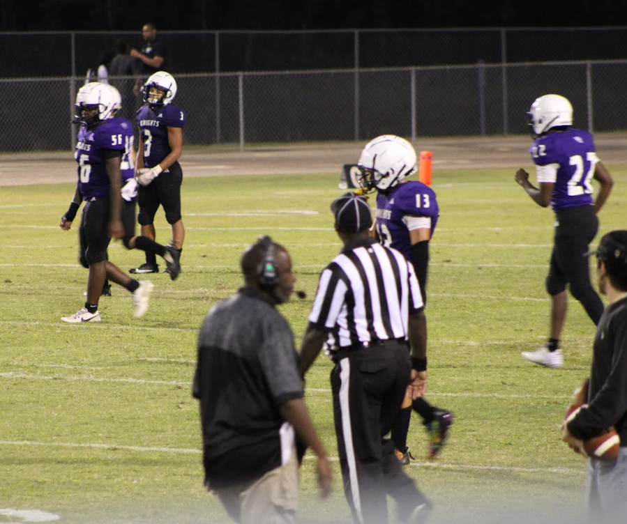 "West Bladen High School junior varsity football players in purple jerseys and white helmets walk across the field during a night game, with referees and coaches visible on the sideline."