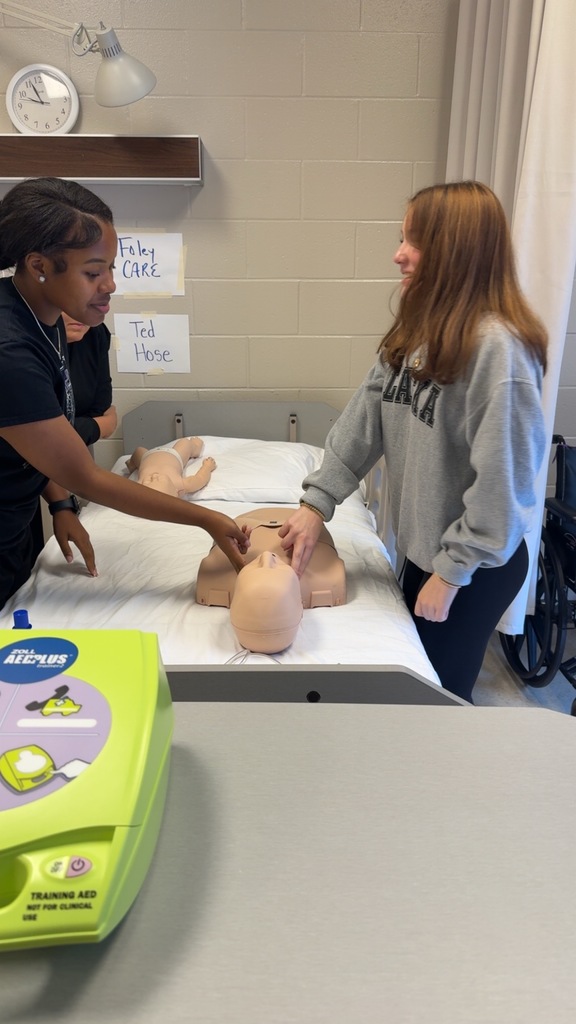 Two students practice CPR on a medical mannequin in a classroom, one giving compressions and the other using a bag valve mask while an instructor observes.