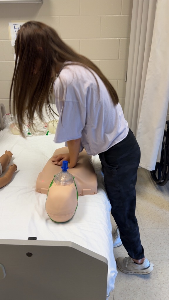 A student presses down firmly on a CPR mannequin’s chest while practicing compressions in a lab setting, with medical supplies nearby.
