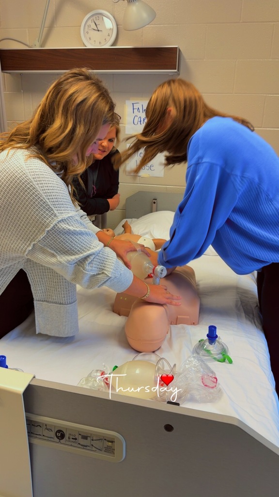 Two students stand by a hospital bed working with a CPR mannequin, one practicing chest compressions and the other locating the pulse, while a training AED is visible in the foreground.