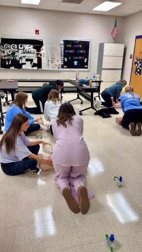 A group of students kneel in pairs on the classroom floor practicing CPR techniques on mannequins, with a bulletin board in the background that reads “We Love Nurse Aides a Latte.”