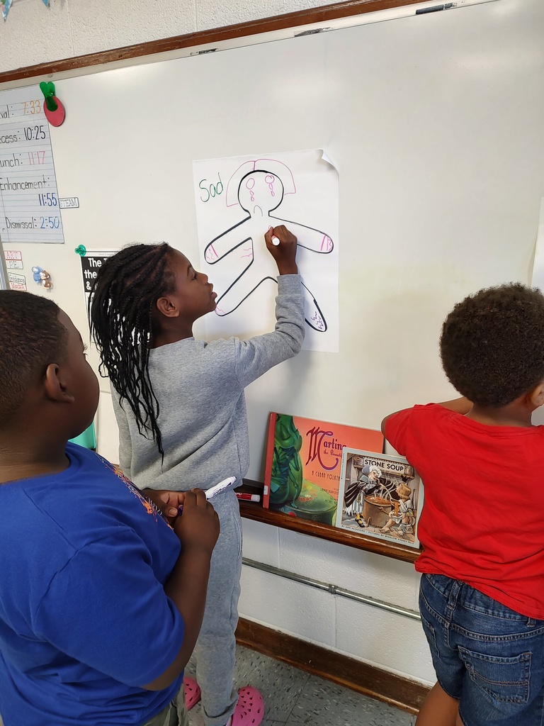A student in gray draws on a large paper figure labeled “Sad,” showing tears and a frown, while two classmates observe and discuss the illustration.