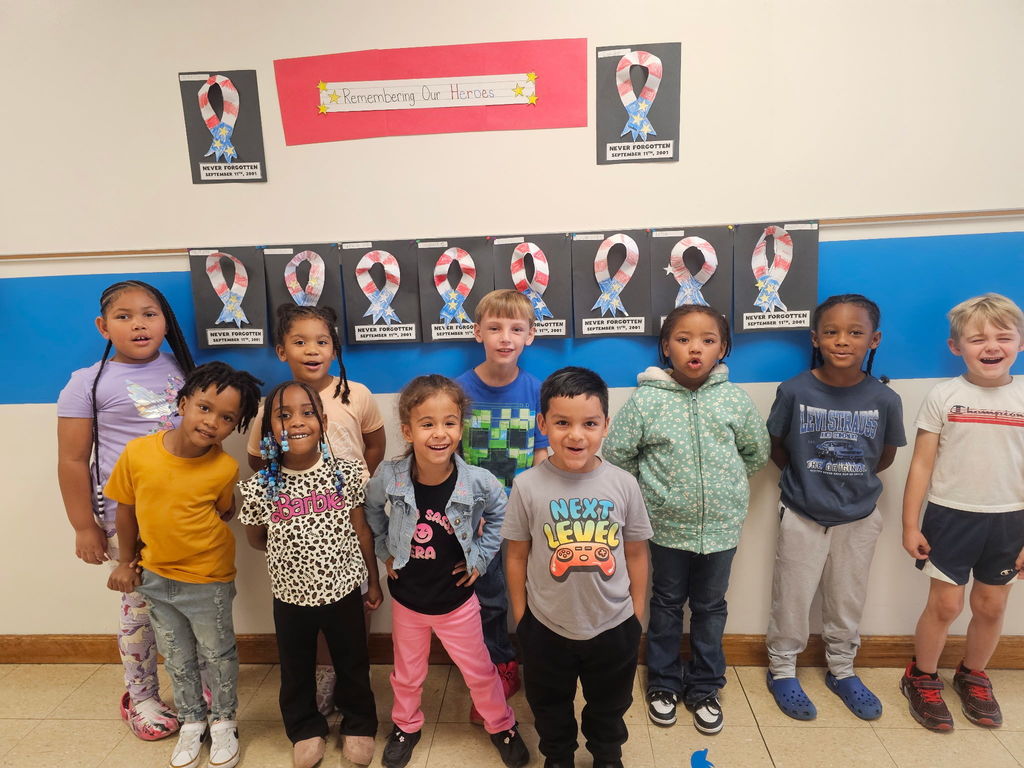 A group of kindergarten students stands smiling in front of a bulletin board display for September 11th. Behind them are red, white, and blue ribbon crafts on black paper, each labeled “Never Forgotten, September 11th, 2001.” At the top of the display is a red banner that reads “Remembering Our Heroes” with yellow stars on each side. The children are standing in a row, showing pride in their work and honoring the memory of the day.