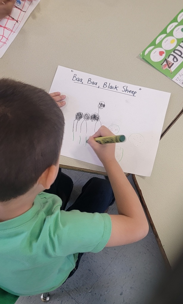 A student in a green shirt colors a picture labeled “Baa Baa Black Sheep,” showing sheep and circular shapes in black and green crayon.