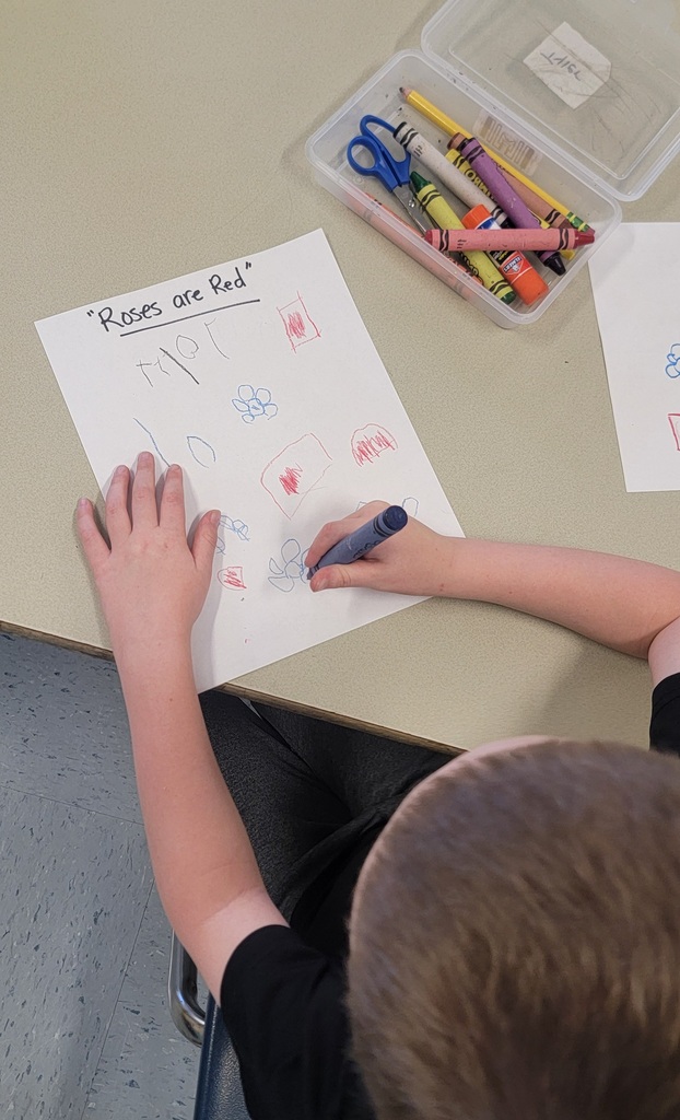 A student draws small shapes and flowers on paper labeled “Roses are Red” while using a blue crayon.