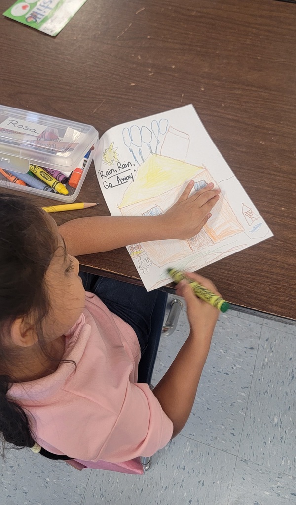 A young student colors a drawing of a house with raindrops, labeled “Rain Rain Go Away,” using crayons at a classroom table.