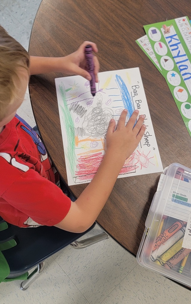 A student in a red shirt works on a colorful page labeled “Baa Baa Black Sheep,” filling it with crayon scribbles and shapes.