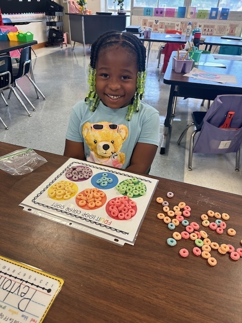 A smiling young girl with green beads in her braids sits at a classroom table. In front of her is a math sorting sheet with colorful cereal pieces grouped by color.