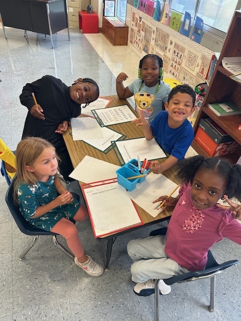 A group of five children sit around a table smiling. They have pencils and papers in front of them, working on a classroom activity together.