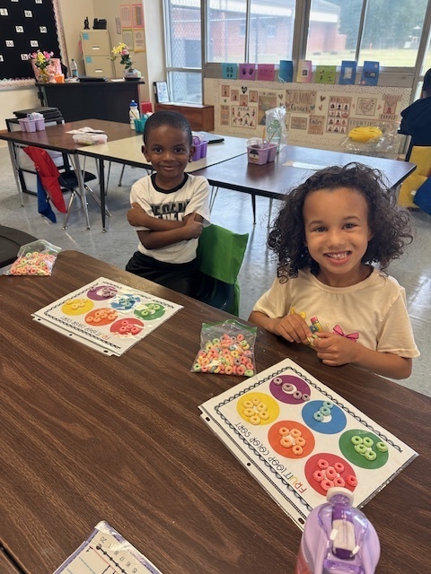 Two children sit together at a classroom table, each with a sorting mat and bag of cereal. They are smiling and showing their work.