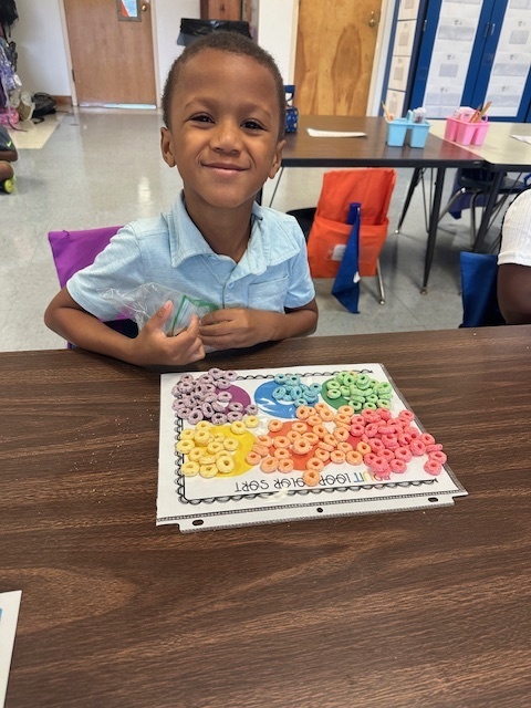 A young boy smiles while holding a bag of cereal. He has sorted colorful cereal pieces onto a worksheet by color at his desk.