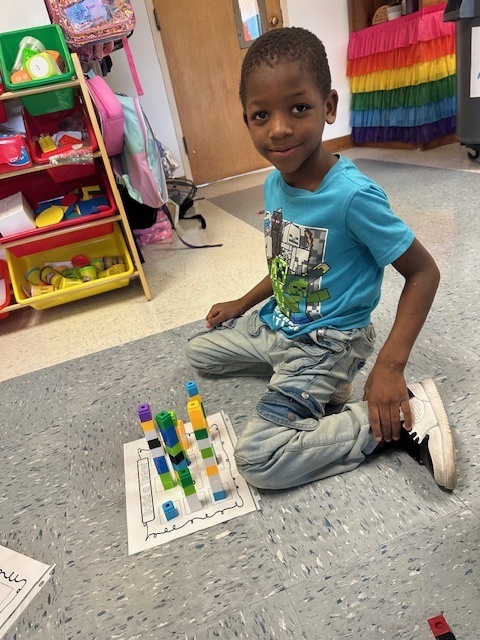 A boy in a blue shirt and jeans sits on the classroom floor with a worksheet and several tall towers of connecting cubes.