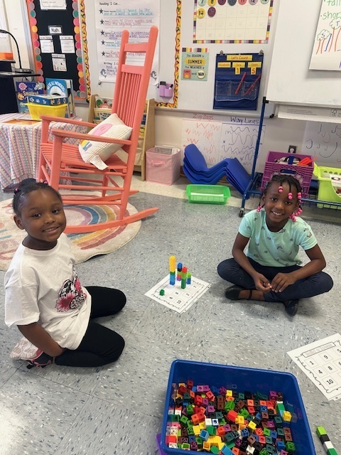 Two girls sit on the classroom floor working with colorful linking cubes. One girl has stacked cubes into tall towers on her paper.