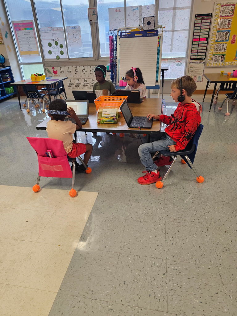 Four students sit at a classroom table, each working on a laptop. They wear headphones and focus on their screens during a technology activity.