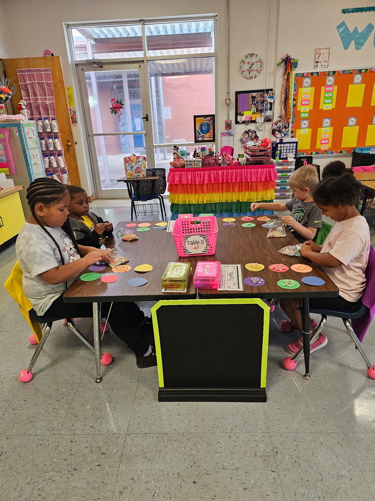Four students sit at a table sorting colorful cereal pieces onto large paper circles by color. The classroom is bright and decorated with rainbow-colored items.
