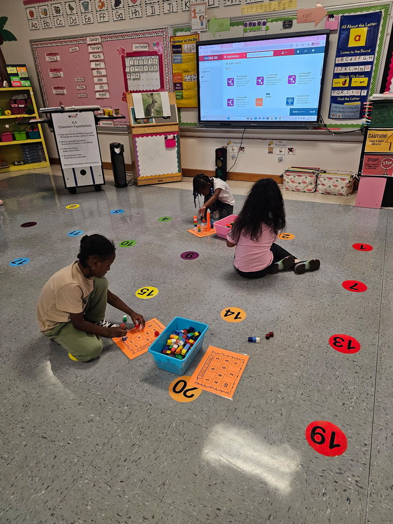 Three students sit on the classroom floor working with colorful connecting cubes and orange math worksheets. Number circles are taped on the floor, and a smartboard displays a lesson at the front.