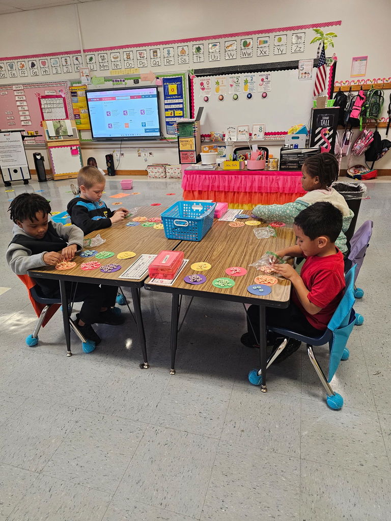 Four students work at a table, carefully sorting cereal pieces onto colored paper circles. The smartboard and alphabet border are visible in the background.