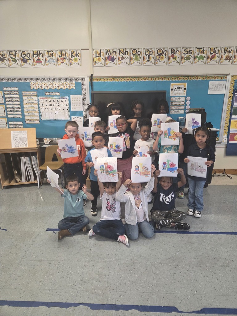 Alt text: A group of kindergarten students stand together in a classroom holding up their colorful drawings. The children are smiling and showing their work related to Habit One: Be Proactive. The background shows alphabet letters across the wall, a classroom calendar, and a bulletin board with bright borders. Some students are kneeling in front while others stand behind them, proudly displaying their completed projects.