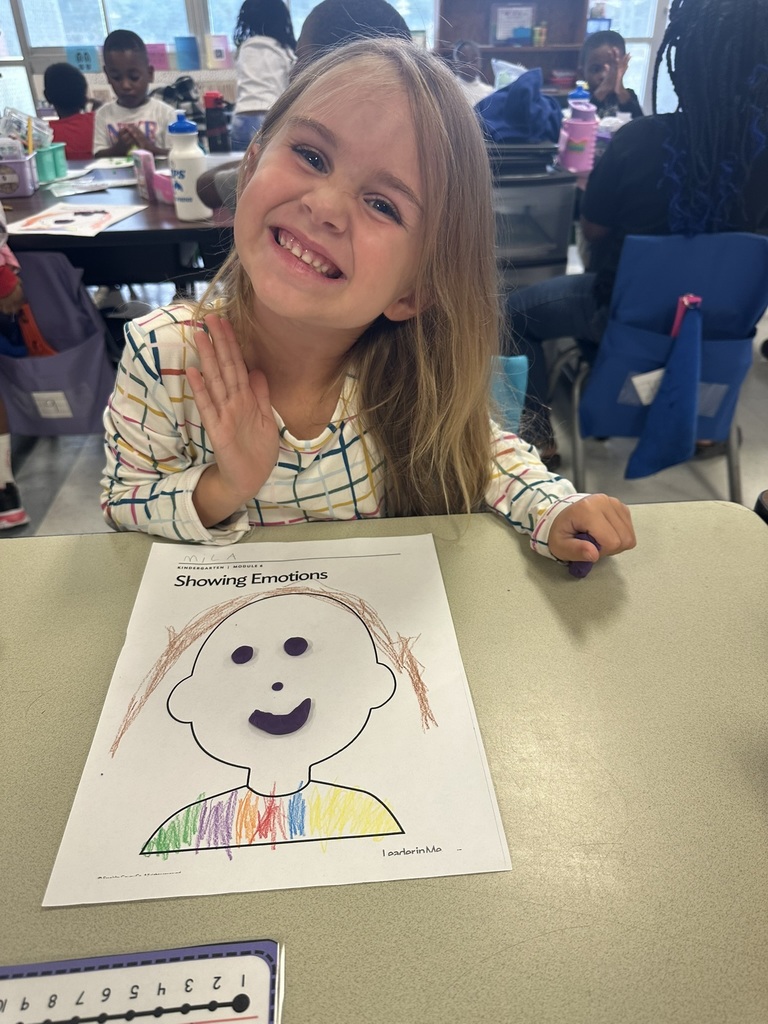 A girl smiles and waves at the camera while sitting at a desk. Her “Showing Emotions” worksheet is colored in bright rainbow shades with Play-Doh facial features.