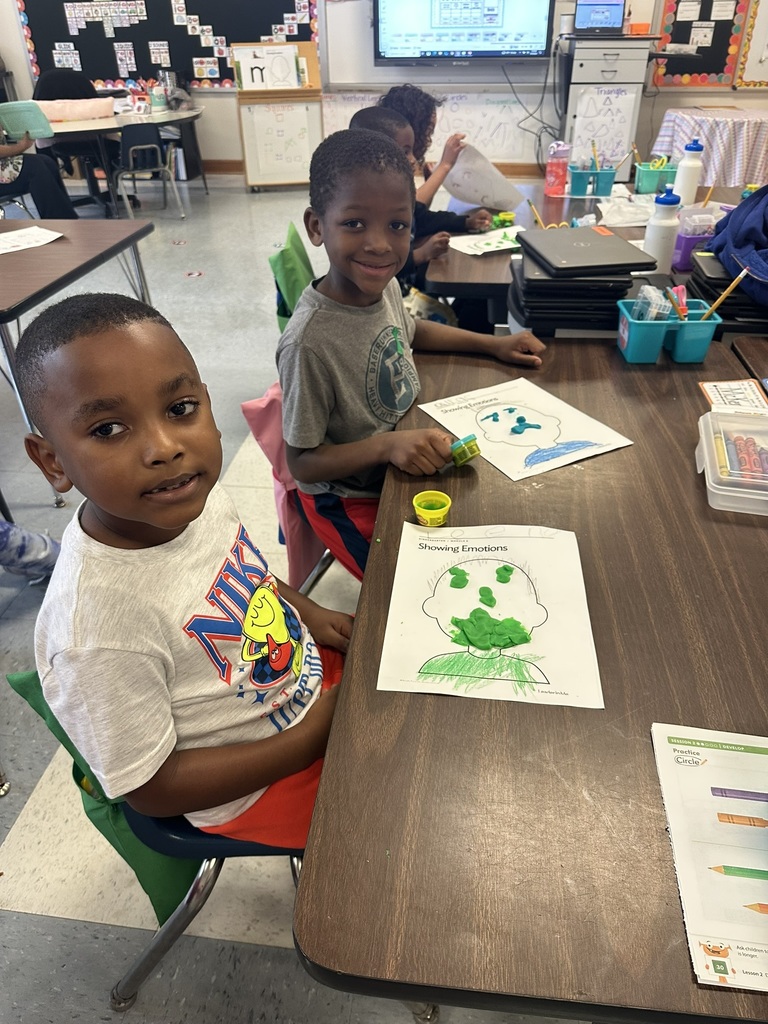 Two boys sit at a classroom table working on “Showing Emotions” worksheets. Their papers are decorated with green Play-Doh faces and bright crayon colors.