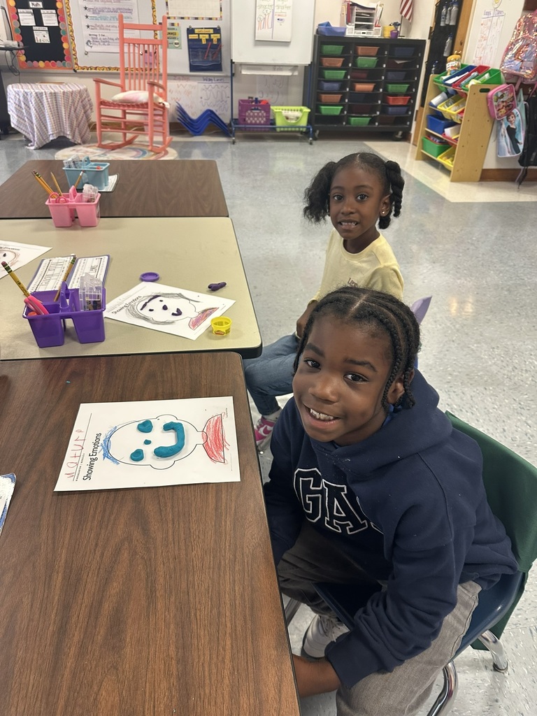 Two young students sit at a classroom table smiling. In front of them are worksheets titled “Showing Emotions,” decorated with colorful drawings and Play-Doh faces.