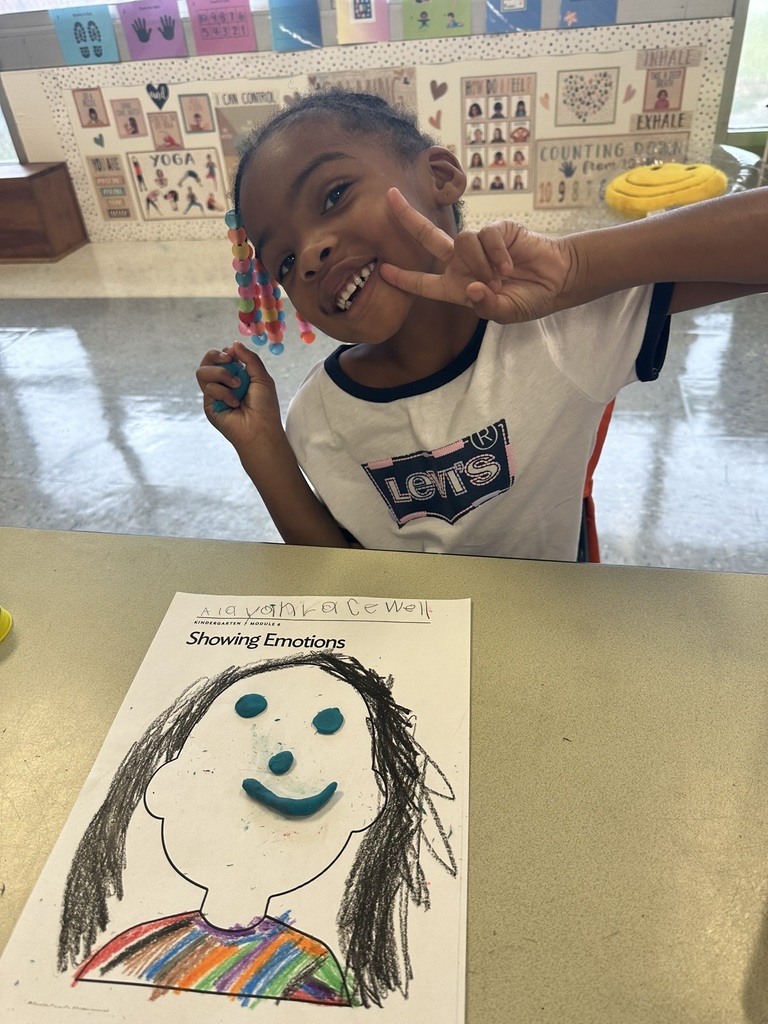 A girl smiles and flashes a peace sign while sitting at her desk. Her “Showing Emotions” worksheet shows a rainbow shirt and a Play-Doh