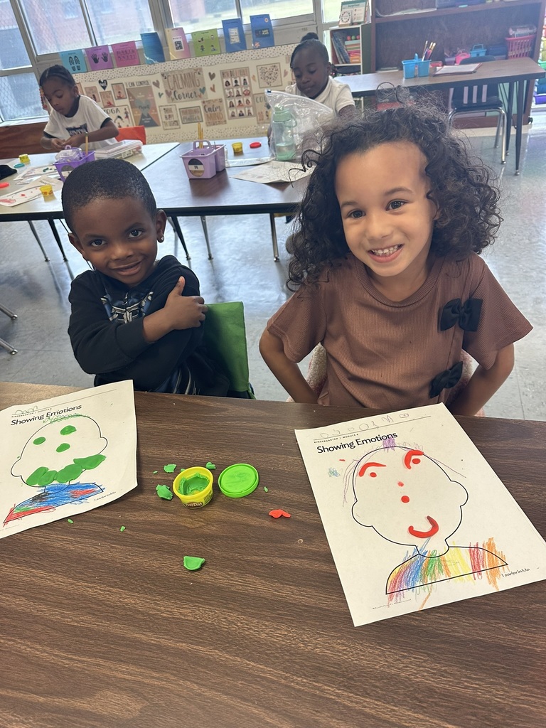 Two students sit side by side at a table, grinning at the camera. Their “Showing Emotions” worksheets are decorated with Play-Doh pieces and bright crayon colors.