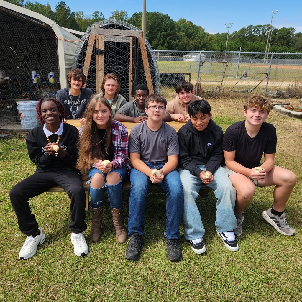 Group of nine students sitting on a bench outside near a chicken coop, smiling and holding baby chicks. A baseball field and trees are in the background