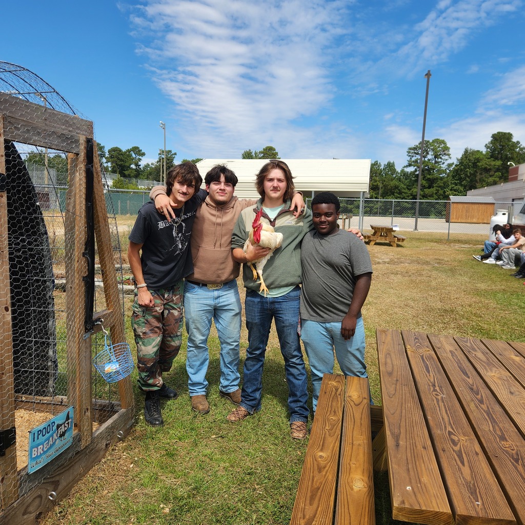 Four students standing together outdoors beside a chicken coop. One student is holding a rooster while the others stand close with arms around each other. Picnic tables and more students are visible in the background.