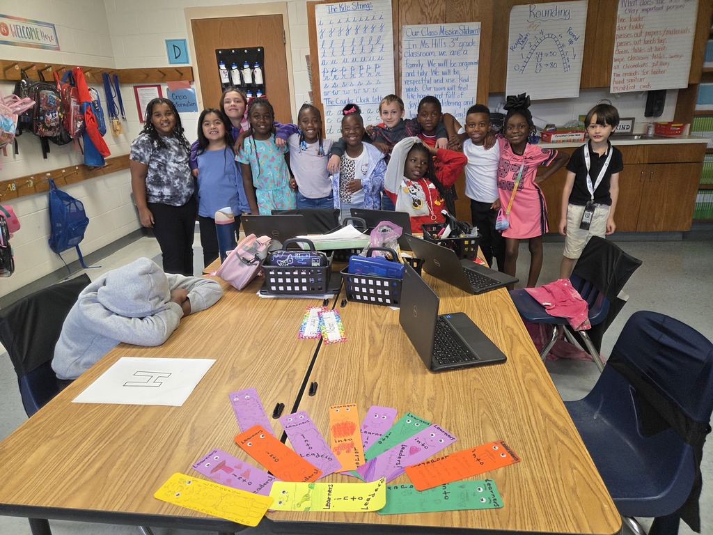 A group of children and one adult stand posing for a photo in a classroom. Laptops, colorful bookmarks, and supplies are on a table in the foreground.