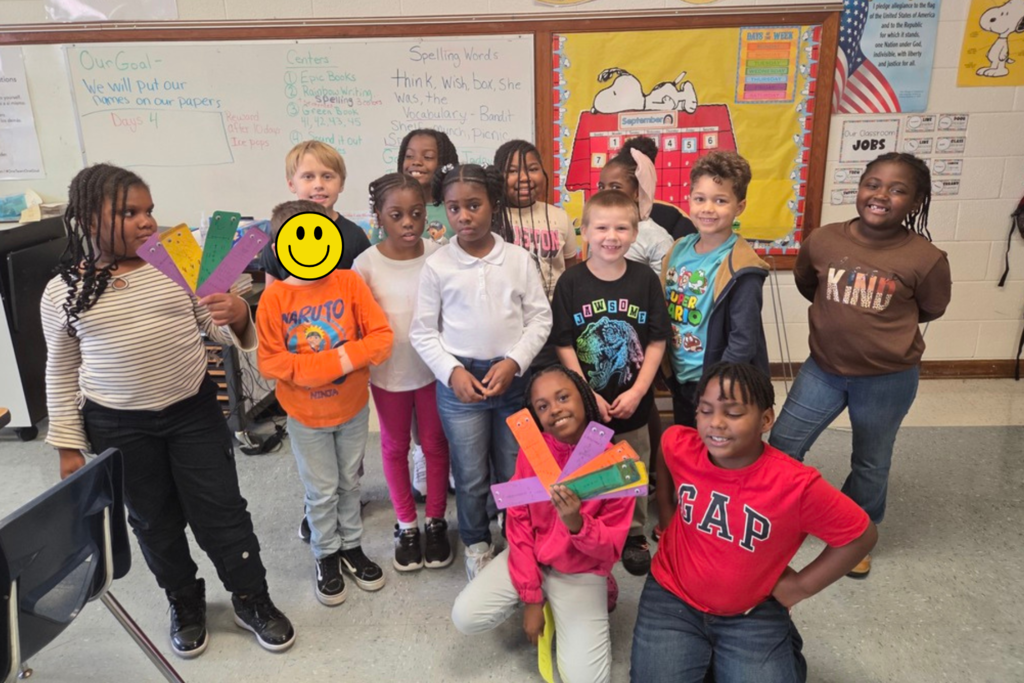 A group of elementary school children stand together in a classroom, posing for a photo in front of colorful educational posters and a bulletin board.