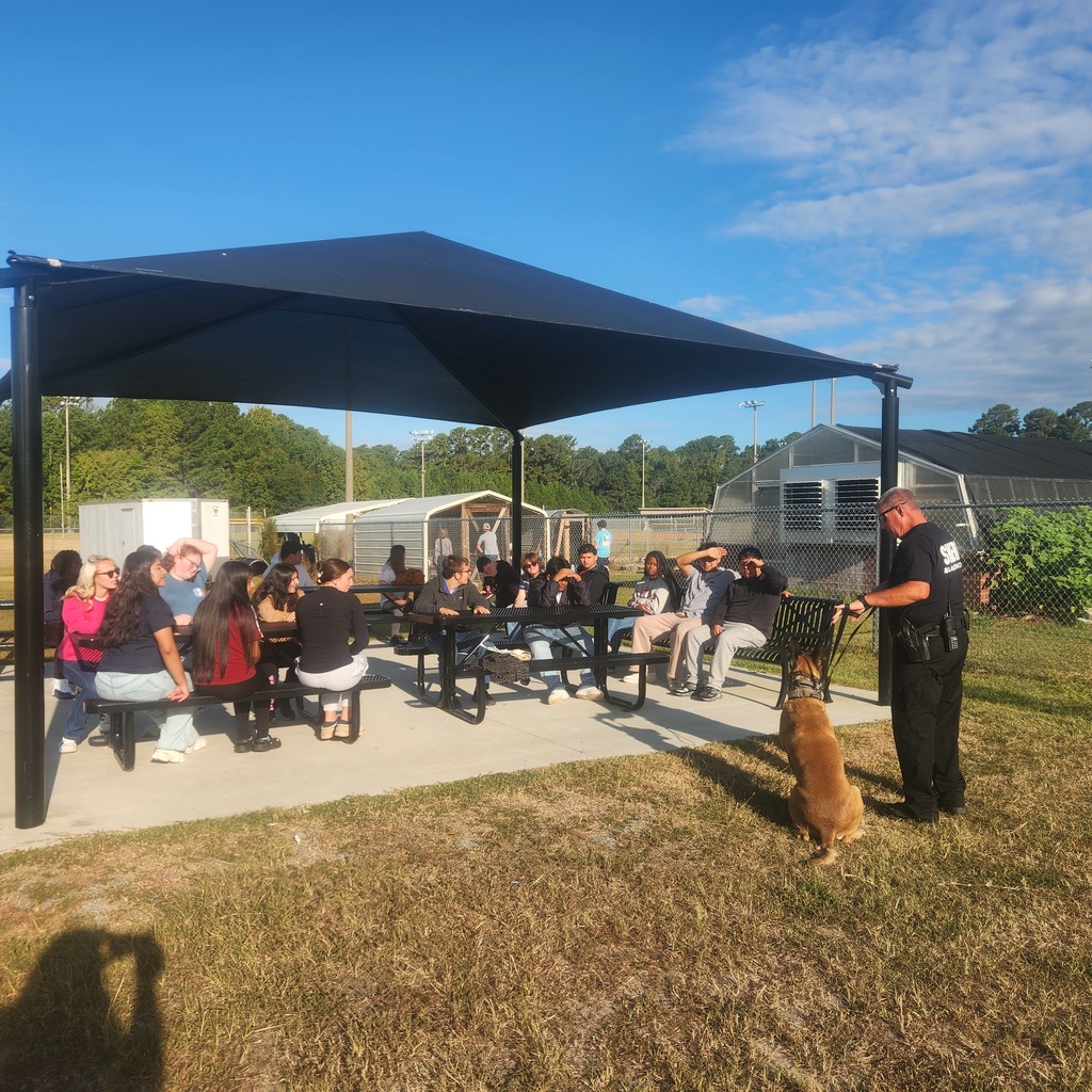 Group of students sitting at picnic tables under a large outdoor canopy on school grounds. A uniformed officer stands to the right holding a working dog, addressing the students. Behind them are chain-link fences, sheds, and greenhouses, with trees and a blue sky in the background