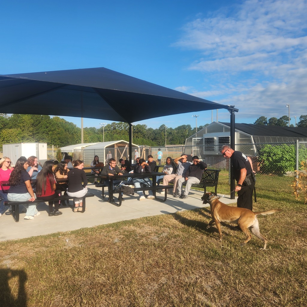 Group of students sitting at picnic tables under a large outdoor canopy on school grounds. A uniformed officer stands to the right holding a working dog, addressing the students. Behind them are chain-link fences, sheds, and greenhouses, with trees and a blue sky in the background