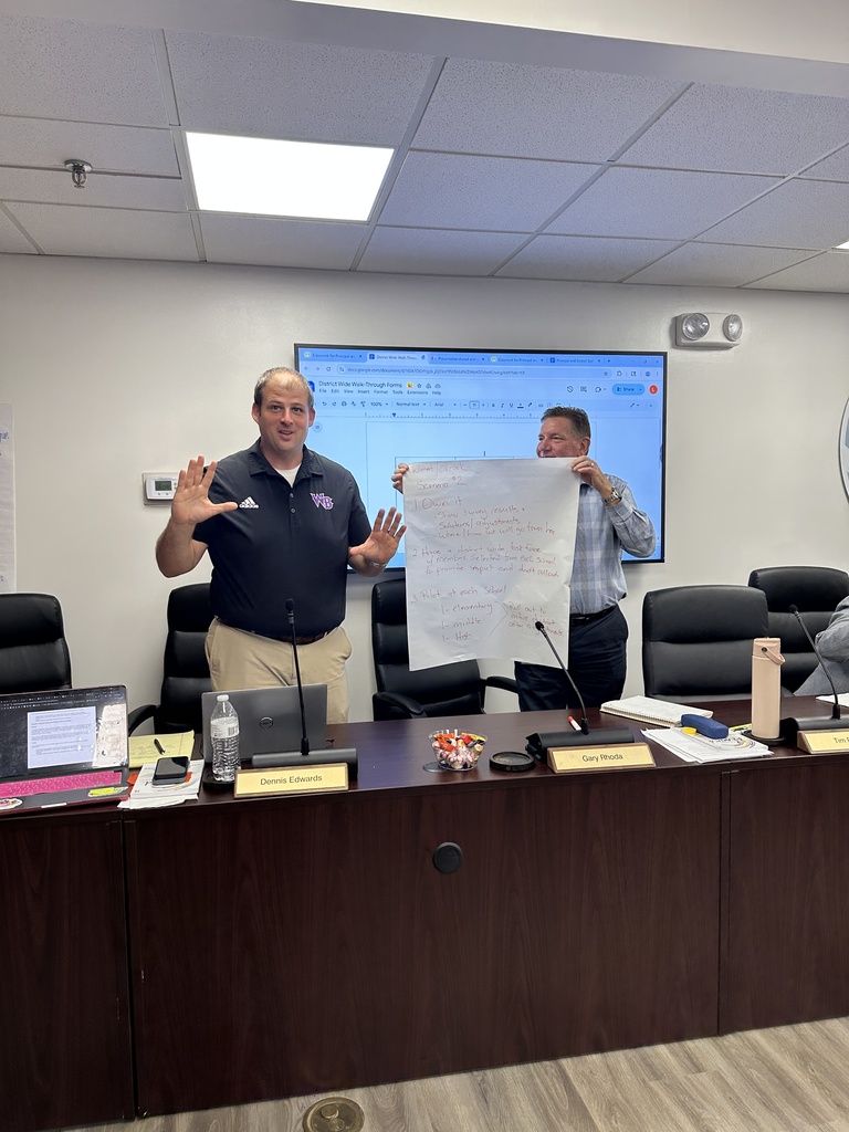 Two men stand at the front of a meeting room, one speaking and gesturing while the other holds up a sheet of chart paper with handwritten notes; laptops and candy are on the table in front of them.