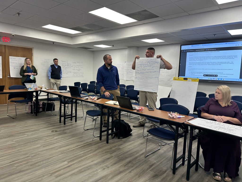 A man in a blue sweater stands holding up chart paper with notes, addressing colleagues in a meeting room; other participants sit and listen, with laptops and papers on the tables.
