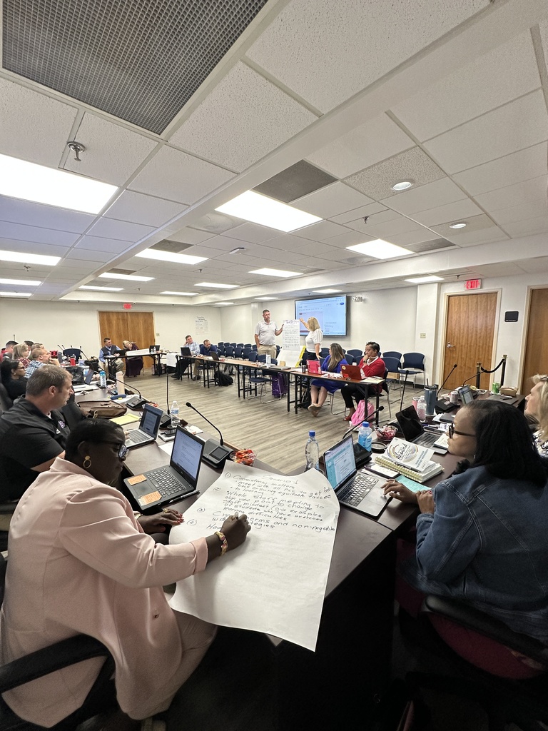 A wide shot of a meeting room where participants are seated around tables; a woman in a pink blazer writes on chart paper in the foreground while others listen and work on laptops.