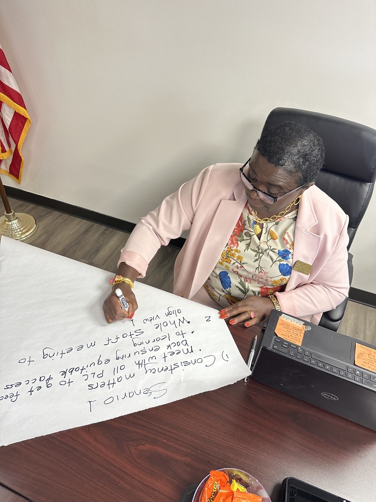 A woman in a pink blazer and floral blouse writes on a large sheet of chart paper during a meeting, with an American flag in the background and a laptop open on the desk beside her.