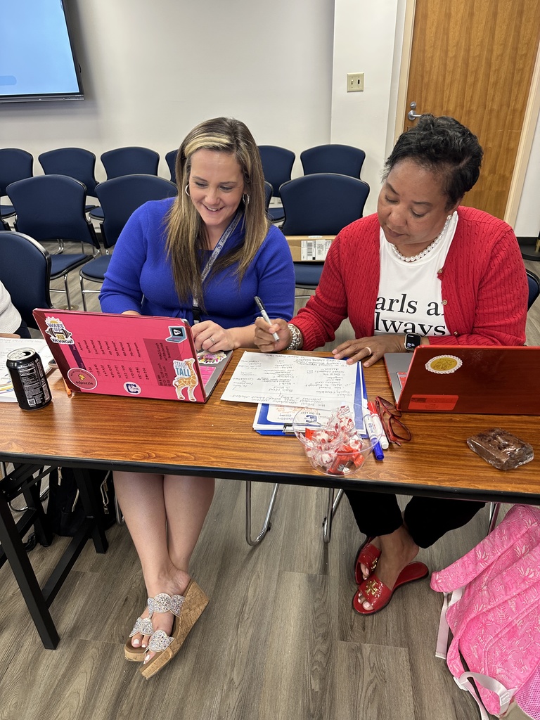 Two women seated side by side collaborate at a table, one smiling while typing on a laptop with stickers and the other writing notes on paper; candy and drinks are on the table.