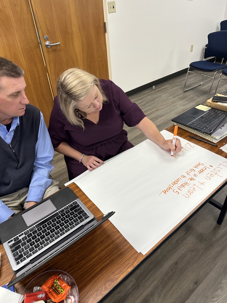 A woman writes notes with an orange marker on chart paper while seated next to a man, both working at a table with laptops and a bowl of candy.