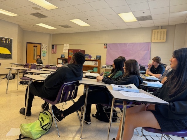 A group of students sit attentively at their desks, listening to classmates present during the mock trial.