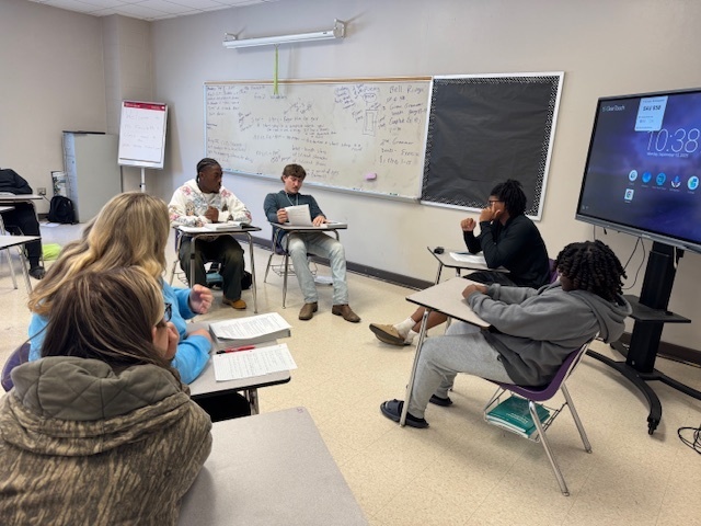 Students sit at desks in a classroom, some in small groups and others individually, preparing notes for the mock trial.