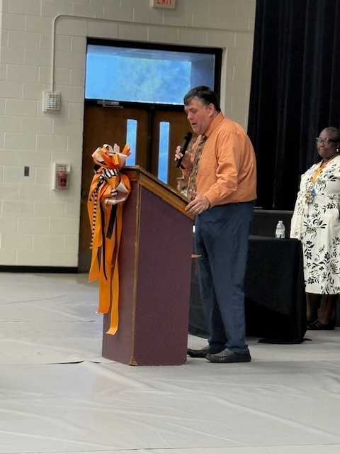 A man in an orange shirt speaks at a podium decorated with orange and black ribbons during the Beta Induction ceremony.