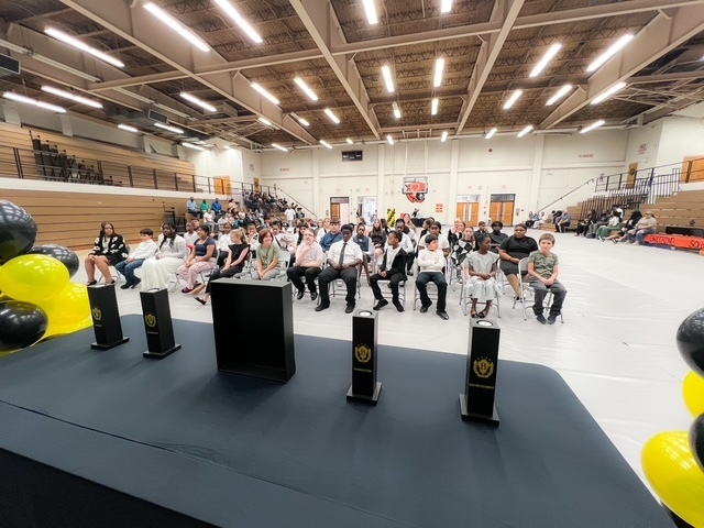 Students and families sit in the gymnasium during the Beta Induction ceremony, viewed from behind the stage decorated with Beta pillars and balloons.