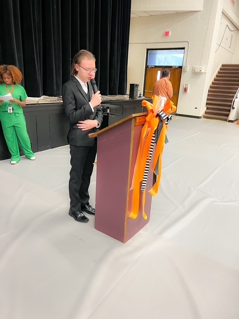 A student in a black suit speaks at a podium decorated with orange and black ribbons during the Beta Induction ceremony.