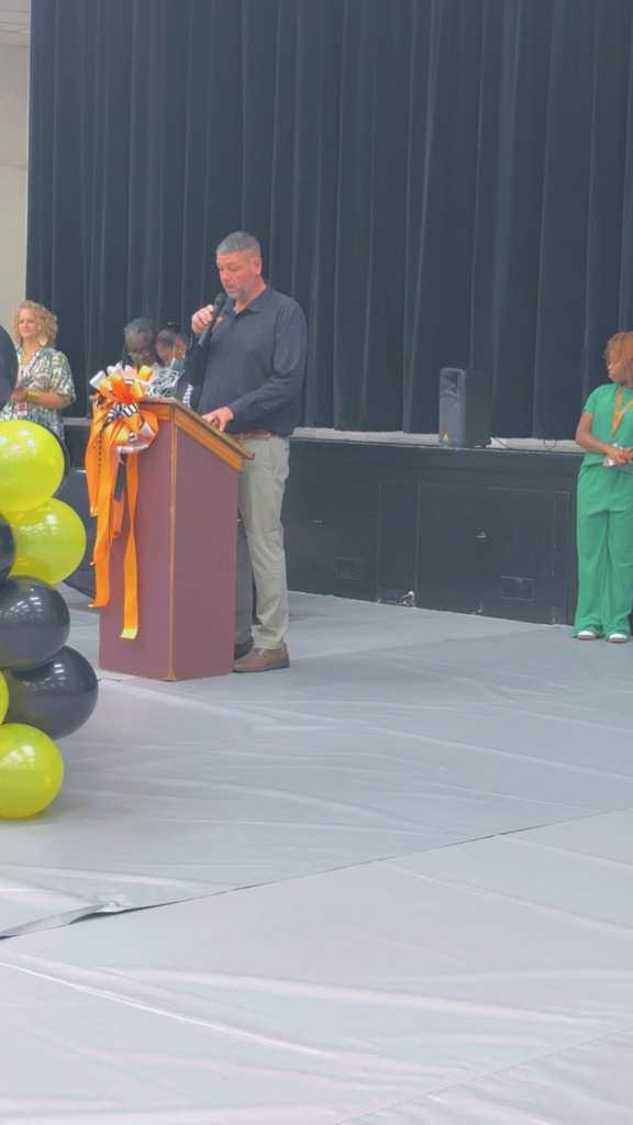 A man in a black shirt speaks at a podium decorated with orange and black ribbons during the Beta Induction ceremony, with black and yellow balloons in the foreground.