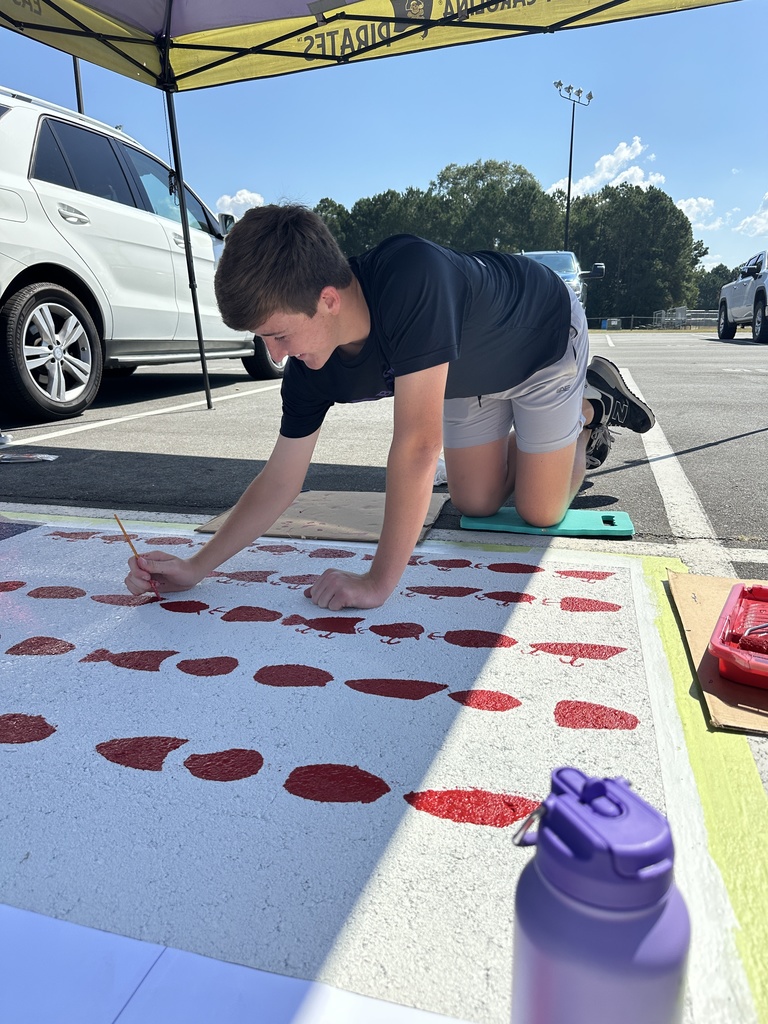 A student kneels under a canopy tent carefully painting red dots across a white parking space design.