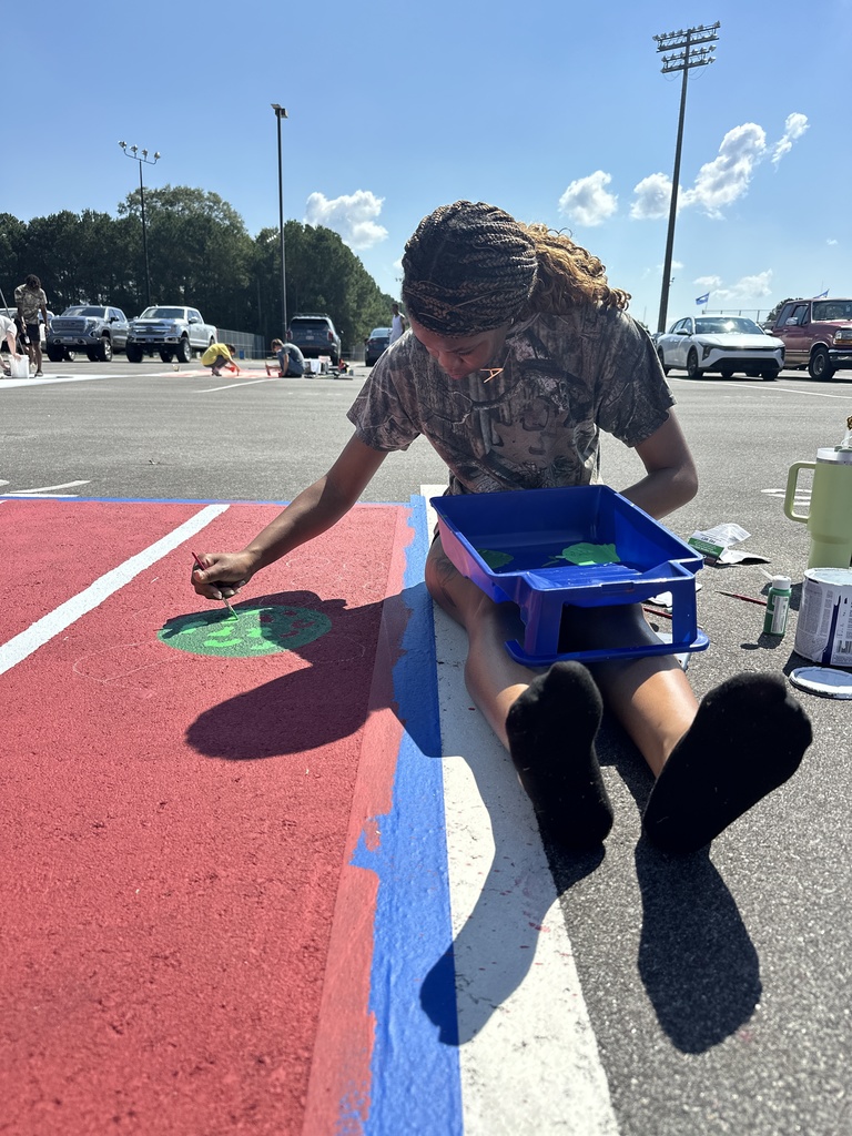 A student sits on the ground painting a small green design on a red parking spot, balancing a paint tray in her lap.