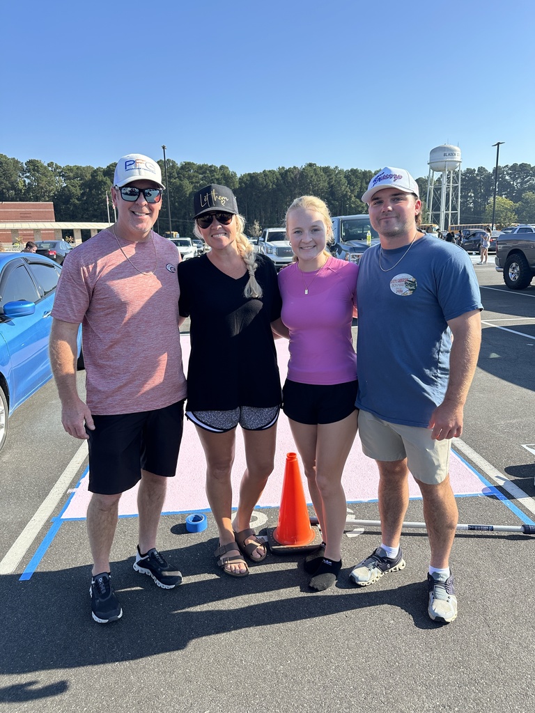 A family of four stands smiling in a parking lot in front of a painted spot, wearing casual summer clothes and hats.